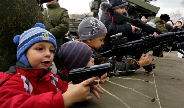 Footage of Ukrainian children learning how to operate military guns, a few out of many people of all ages doing the same.